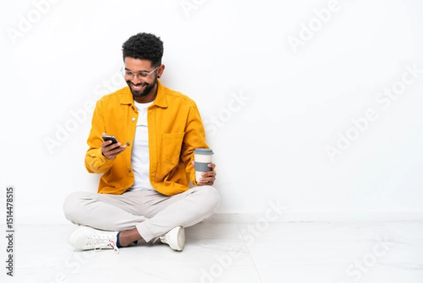 Fototapeta Young Brazilian man sitting on the floor isolated on white background holding coffee to take away and a mobile
