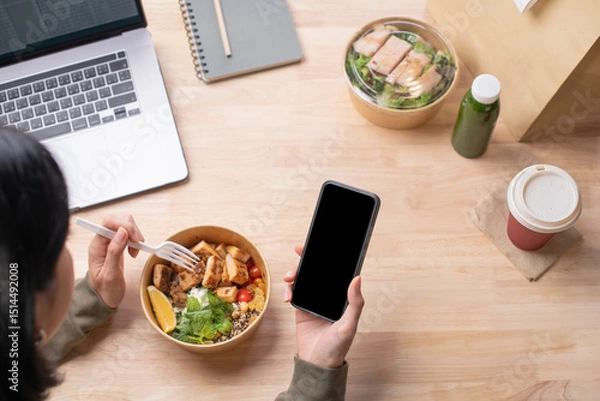 Fototapeta Asian woman eating healthy meal while working, Businesswoman having a lunch break at the office, hand holding phone with blank black screen for copyspace