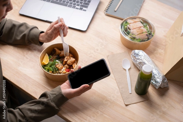 Fototapeta Asian woman eating healthy meal while working, Businesswoman having a lunch break at the office, hand holding phone with blank black screen for copyspace