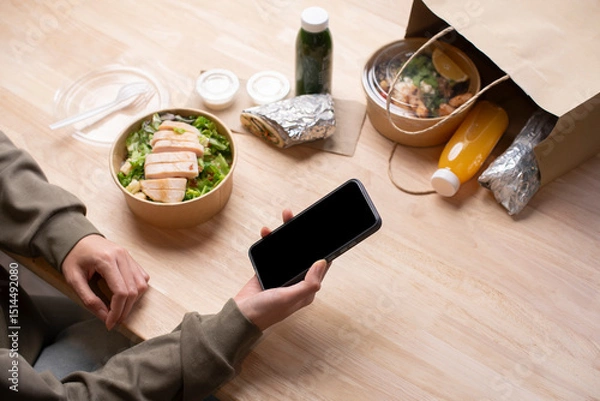 Fototapeta Asian woman eating healthy meal while working, Businesswoman having a lunch break at the office, hand holding phone with blank black screen for copyspace