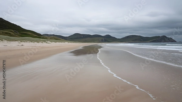 Fototapeta Empty beach with gentle waves and moody sky, soft sand stretching along shoreline with distant green hills under cloudy weather creating calm and peaceful atmosphere
