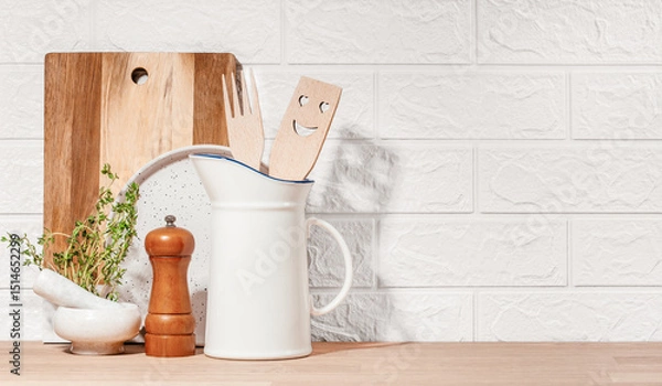 Obraz Kitchen utensils including a smiling wooden spatula, pepper mill, mortar with thyme, and cutting board arranged on a wooden countertop against a white brick wall for a cozy home setting