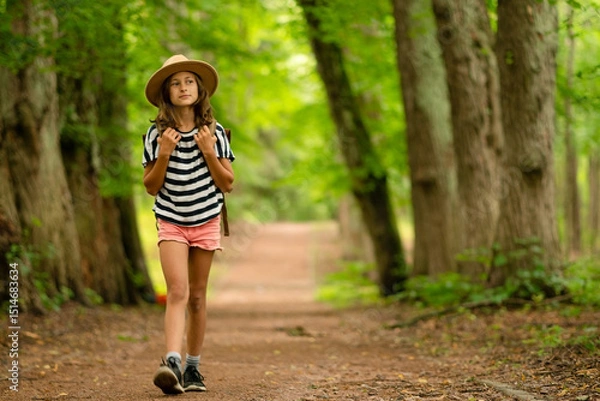 Obraz A full-body view of a dreamy girl walking with a backpack and hat, observing the forest around her. Peaceful summer hiking trip full of curiosity, nature, and exploration.