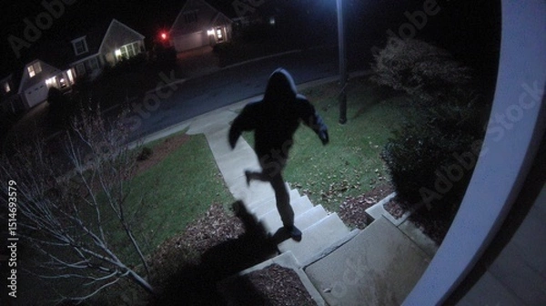 Obraz A person in a hoodie approaches a house at night, caught by a security camera on a dimly lit suburban street.