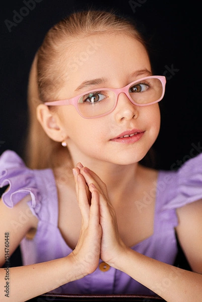 Fototapeta Close-up portrait of a charming young girl in pink glasses and a purple frilly dress posing on a black background. She has her palms folded together. Her hair is tied in a tight ponytail at the back.