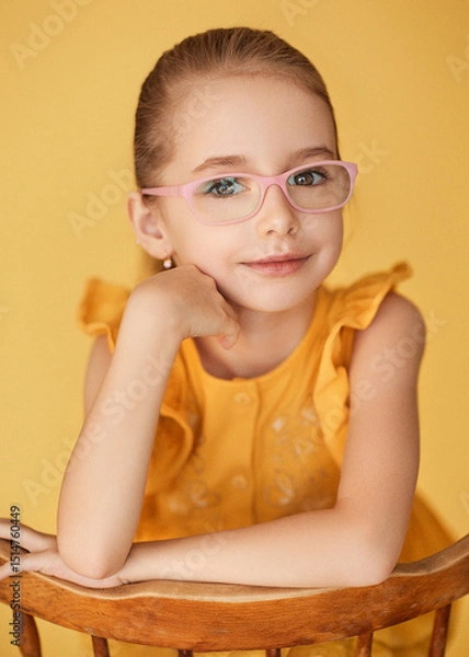 Fototapeta Close up of young girl with blonde hair pulled back wearing yellow ruffled top. She sits leaning on her arm and smiles tenderly. The model with expressive eyes wears corrective glasses with pink frame