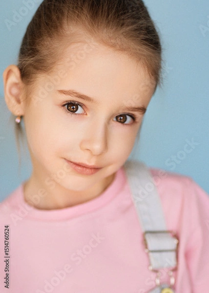 Fototapeta Close-up portrait of a cute girl with light brown hair combed back, wearing a pink shirt and light overalls. She has a gentle expression. The background is soft blue, creating a calm atmosphere.
