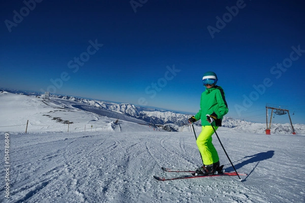 Obraz Skier in green jacket stands upright on skis overlooking valley