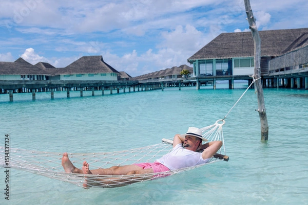 Fototapeta A young man in summer clothes and a hat is lying in a hammock in clear sea water. A carefree man is relaxing by the ocean. Bungalows for tourists in the background.