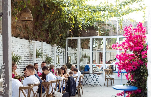 Fototapeta A festive banquet outdoors in the garden. Elegant guests at the tables. Rustic stylish interior. Grapes on the walls and flowering trees.