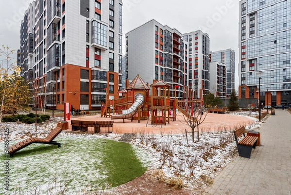 Fototapeta Play area with a wooden complex and benches in the courtyard of multi-story buildings, the ground is covered with light first snow.
