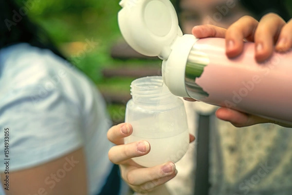 Fototapeta woman pouring warm water from a thermos into a baby bottle to prepare infant formula during a park outing