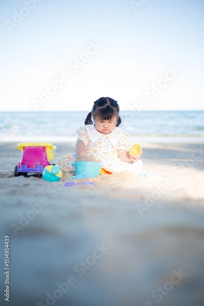 Fototapeta A little girl is playing in the sand on the beach,Happy fun Asian child cute little girl playing sand with toy sand tools at a tropical sea beach in holiday summer on sunset time, tourist trip concept