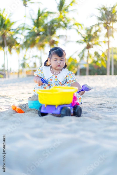 Fototapeta A little girl is playing in the sand on the beach,Happy fun Asian child cute little girl playing sand with toy sand tools at a tropical sea beach in holiday summer on sunset time, tourist trip concept