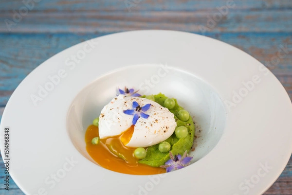 Obraz A fresh pea puree is beautifully plated, featuring a poached egg at the center and adorned with borage flowers. The dish showcases vibrant colors and textures, perfect for any spring menu.