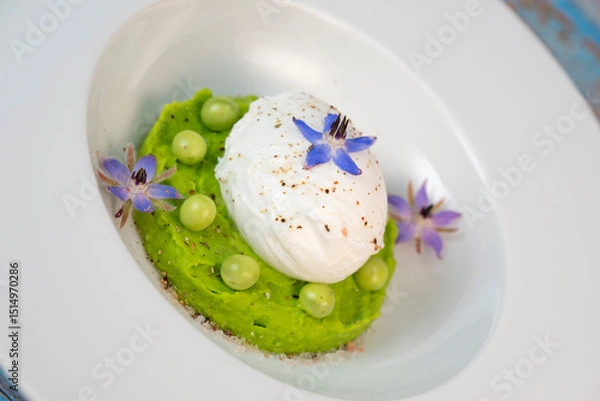 Obraz A fresh pea puree is beautifully plated, featuring a poached egg at the center and adorned with borage flowers. The dish showcases vibrant colors and textures, perfect for any spring menu.