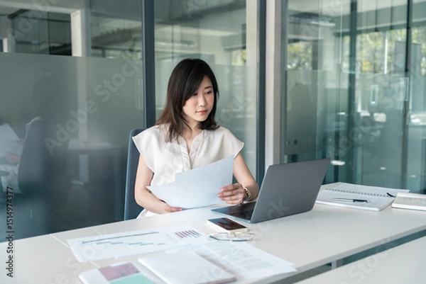 Fototapeta Busy Asian accountant business woman working on laptop computer reading financial document report in office. Middle-aged entrepreneur bank manager businesswoman doing finance paperwork work using pc
