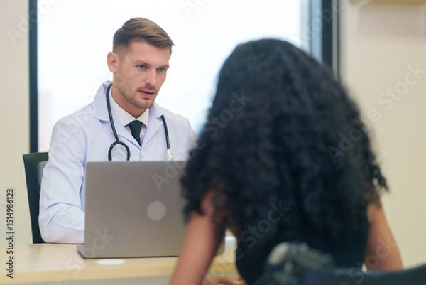 Obraz A caucasian doctor in white gown with stethoscope and laptop on table talking to a woman disabled person patient on wheelchair in the examine room