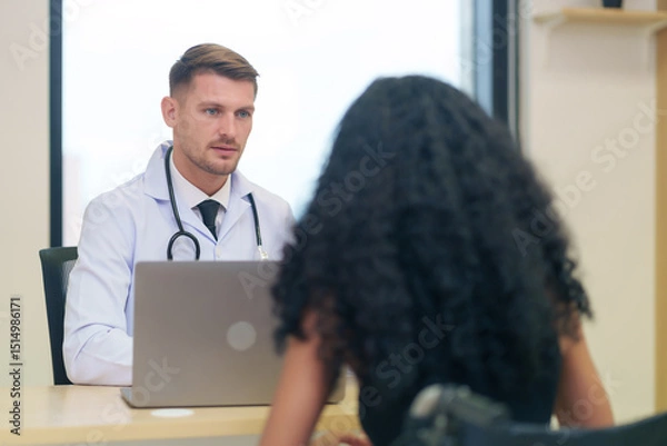 Obraz A caucasian doctor in white gown with stethoscope and laptop on table talking to a woman disabled person patient on wheelchair in the examine room