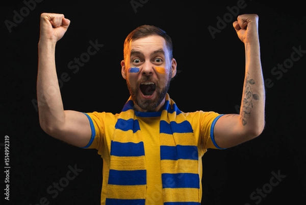 Fototapeta Portrait of euphoric soccer fan man in yellow-blue t-shirt and scarf and with painted face, cheering for his favourite team, making winner's gesture raising his hands to the top and clenching fists