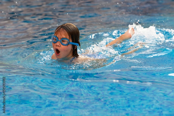 Fototapeta Joyful girl in swim goggles bursts from pool water. A vivid summer scene capturing childhood fun, sport, and games in the pool.