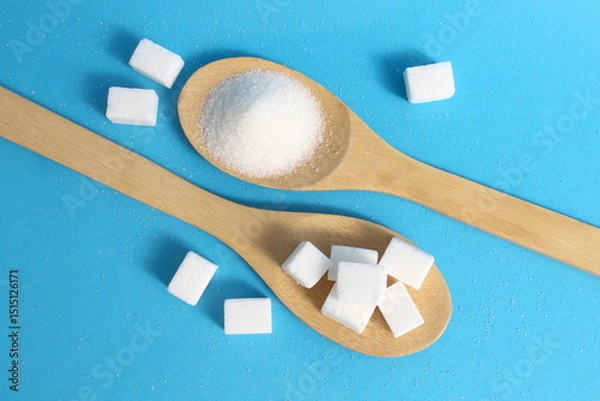 Fototapeta On a blue background, on wooden spoons, there are two types of sugar, loose and cubed.