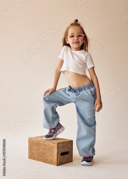 Fototapeta A little mischievous girl with glasses stands in a casual pose with one foot on a wooden box. She's wearing a white crop top and wide jeans. The model with blonde hair poses in a studio on a cyclorama