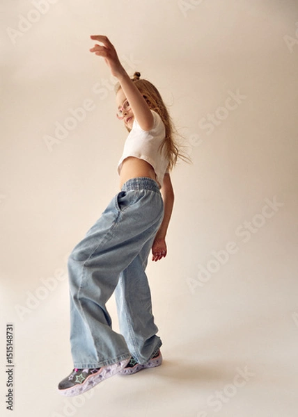 Fototapeta An energetic young girl stands in a dynamic pose, raising one arm up. Her long blonde hair flies to the sides. The child in a white crop top and jeans tries to keep her balance, standing on one leg.