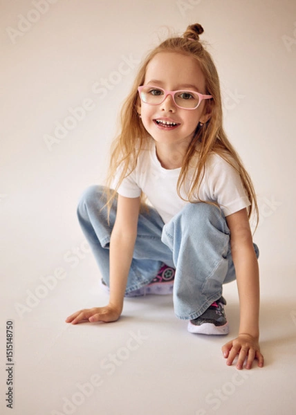 Fototapeta An active and cheerful young girl with long blond hair and glasses sits on her haunches and smiles. She touches the floor with her hands. A child in a white T-shirt and blue jeans poses on a cyclorama