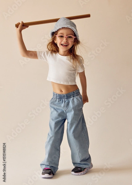 Fototapeta A little girl in a white T-shirt and wide jeans holds a wooden stick above her head and laughs happily. She wears a blue Panama hat, pink glasses. She is playing with a bat joyfully and energetically.