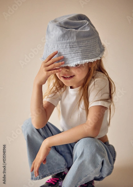 Fototapeta A cute seven-year-old child in a light blue headdress that partially covers her eyes is squatting. The little girl in a white T-shirt and jeans playfully touches the brim of her hat with her hand.