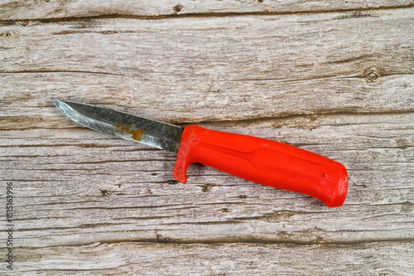 Obraz A red-handled knife lies on a rustic wooden surface, showcasing signs of use and wear. The blade features a worn edge, reflecting its practical purposes over time