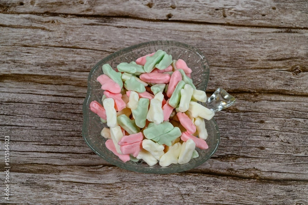 Obraz Brightly colored candy pieces in pastel shades are arranged in a crystal dish on a weathered wooden table. They create a cheerful and inviting display ideal for snacking or sharing