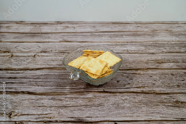 Obraz A clear glass bowl holds an assortment of crackers arranged neatly on a rustic wooden surface. The setting suggests a casual snack time at home, perfect for entertaining