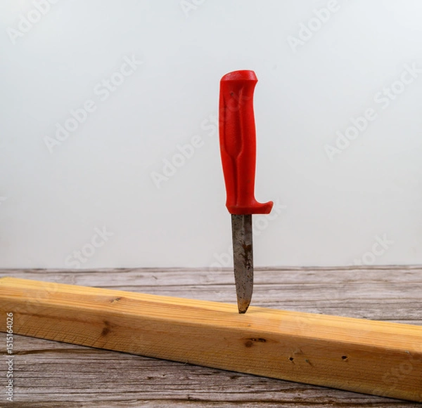 Obraz A knife with a bright red handle is embedded firmly into a wooden plank resting on a rustic table. The background is light and neutral, enhancing the focus on the knife