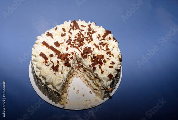 Obraz A creamy cake topped with whipped frosting and chocolate shavings sits on a white plate. A slice is cut, indicating it's ready to be enjoyed at a festive gathering