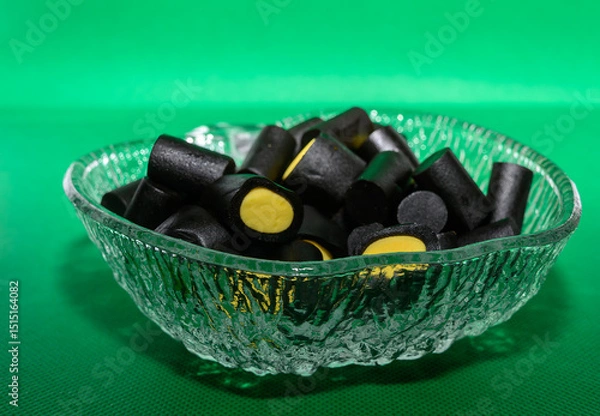 Obraz A glass bowl filled with black licorice candies featuring vibrant yellow centers is displayed against a vivid green background. These treats bring a playful touch to the gathering