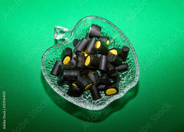 Obraz A glass bowl filled with black licorice candies featuring vibrant yellow centers is displayed against a vivid green background. These treats bring a playful touch to the gathering