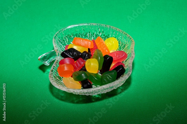 Obraz A clear decorative bowl holds a variety of colorful jelly candies, including orange, yellow, green, and red. The vibrant colors contrast against a bright green backdrop
