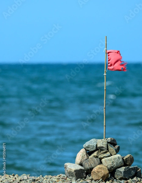 Obraz A tattered red flag is perched on a stick, resting atop a pile of stones near calm blue waters. The clear sky enhances the serene coastal atmosphere during daylight