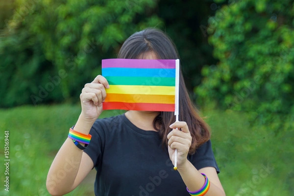 Fototapeta An Asian LGBT woman wearing a black t-shirt and a rainbow wristband holds up a rainbow flag in front of her face as a symbol of LGBT pride.