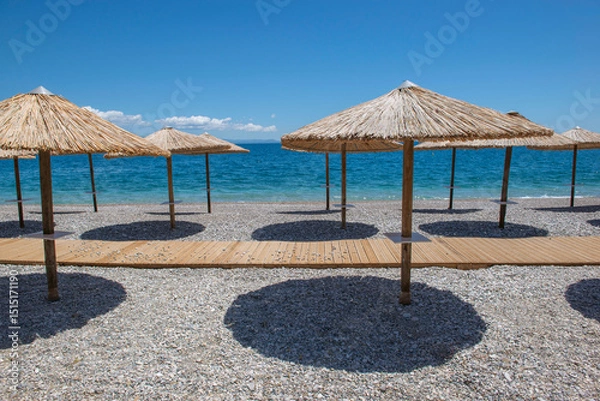 Fototapeta straw umbrella on a beach iof pebbles n Peloponnese, Greece with their well drawn shadow in front beautiful sea