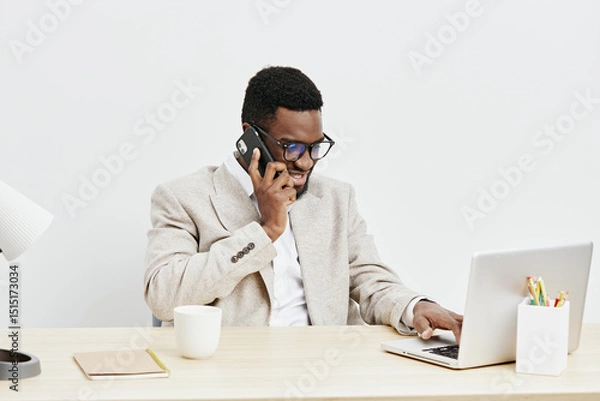 Fototapeta Professional man with dark skin, wearing glasses and beige blazer, talking on the phone while working on a laptop in a modern office space. Business, communication, technology, workspace, people