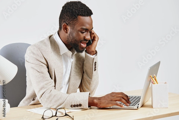 Fototapeta Smiling young man working on a laptop at a desk in a modern office setting, wearing a beige cardigan and white shirt, with glasses and stationery nearby, conveying productivity and professionalism
