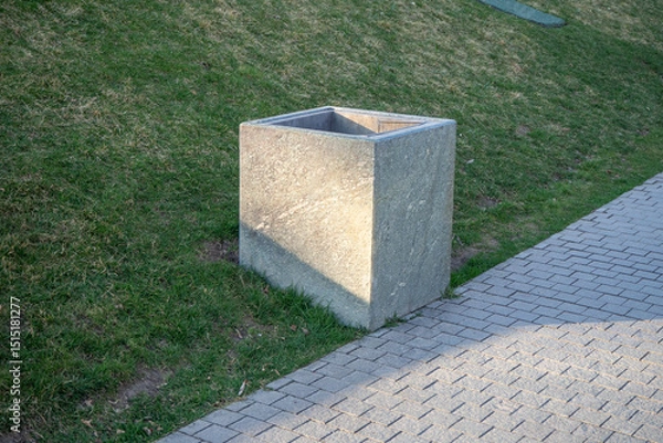 Fototapeta Square Concrete Trash Can on Gras. Concrete outdoor trash can with square shape placed on grass near a stone path. Designed for public park waste disposal. Urban landscape element.