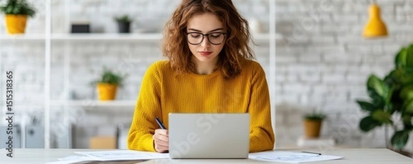 Fototapeta Coworking spaces, shared office and productivity, Young woman in glasses working on a laptop at a desk in a bright, modern home office with plants and shelves in the background.