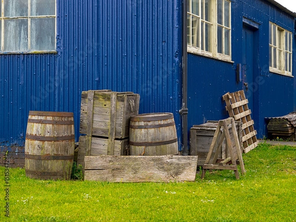Fototapeta Rustic wood barrels and crates beside a weathered blue building on grass.