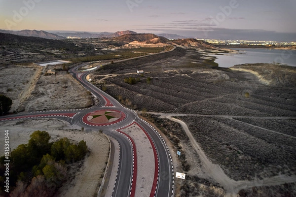Fototapeta Aerial view of curved road and modern roundabout surrounded by dry fields and hills near large reservoir at dusk. Spain