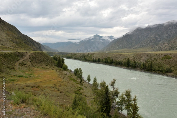 Obraz High banks of a wide mountain river flowing through a long valley surrounded by rocks on a cloudy summer day.