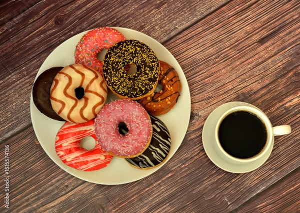 Obraz Cup of black coffee on a saucer and a plate with an assortment of fresh donuts in colorful glaze on a wooden table.